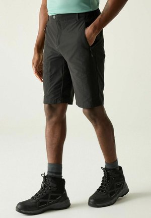 Mann trägt schwarze Shorts mit Reißverschluss, graue Socken und schwarze Wanderschuhe und steht mit einer Hand in der Tasche.