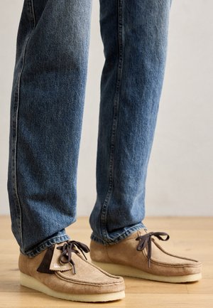 Mocasines de gamuza en color marrón claro con acentos en marrón oscuro, puntera redondeada y suela blanca suave. Presenta un diseño con cordones y detalles de costura.