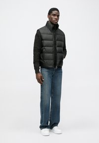 Μαύρο επενδεδυμένο γιλέκο με ψηλό γιακά, σε συνδυασμό με μαύρο υφασμάτινο τοπ από κάτω, μπλε τζιν με ίσια γραμμή και λευκά αθλητικά παπούτσια.