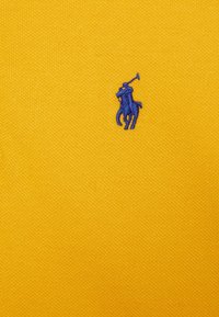 Keltainen kangastausta, jossa on pieni sininen brodeerattu polo-pelaajan logo, esittelee tekstuuripintaista kudontaa.