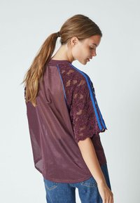 Burgundy mesh shirt with sheer lace sleeves, featuring blue stripes along the shoulders. Loose fit and lightweight texture.