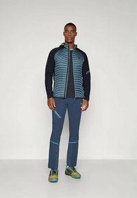 Moška zunanja jakna z teksturiranim temno zelenim zgornjim delom in črnimi rokavi, v kombinaciji s temno modrimi hlačami z zelenimi poudarki ter zelenimi čevlji.