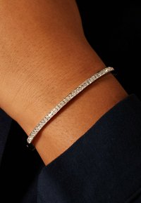 Zilveren armband met een rij heldere stenen langs de bovenzijde, glad oppervlak, gedragen om een pols tegen een donkere stoffen achtergrond.