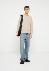 Suéter de canalé beige, jeans de pierna ancha azul claro y calzado negro. Una bolsa de hombro negra reposa en su hombro. Fondo blanco liso.
