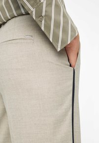 Beige broek met een lichte textuur, voorzien van een zijzak en zwarte bies. Olijfgroen gestreept shirt zichtbaar bij de kraag.