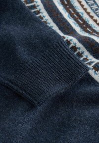 Detail eines marineblauen Strickpullovers mit geripptem Bündchen, weicher Textur und einem gemusterten Joch mit blauen, braunen und weißen Akzenten.