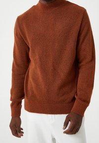 Suéter marrón de punto con cuello alto, mangas largas y puños acanalados, que presenta una textura suave y un ajuste holgado.