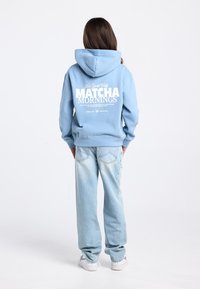 Lichtblauwe hoodie met witte tekst op de achterkant, met de tekst "New York City MATCHA MORNINGS." Gecombineerd met lichtblauwe jeans en witte sneakers.