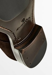 Ruskea mokkanahkakengät, joissa on sileä nahkasuoja, metalliset yksityiskohdat kantapäässä ja näkyvät ompelut yksityiskohdissa sisävuorauksessa.