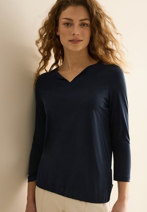 Haut à manches longues bleu marine avec un décolleté en V, texture lisse, ourlet légèrement asymétrique et manches 3/4. Présente une étiquette sur le côté gauche.