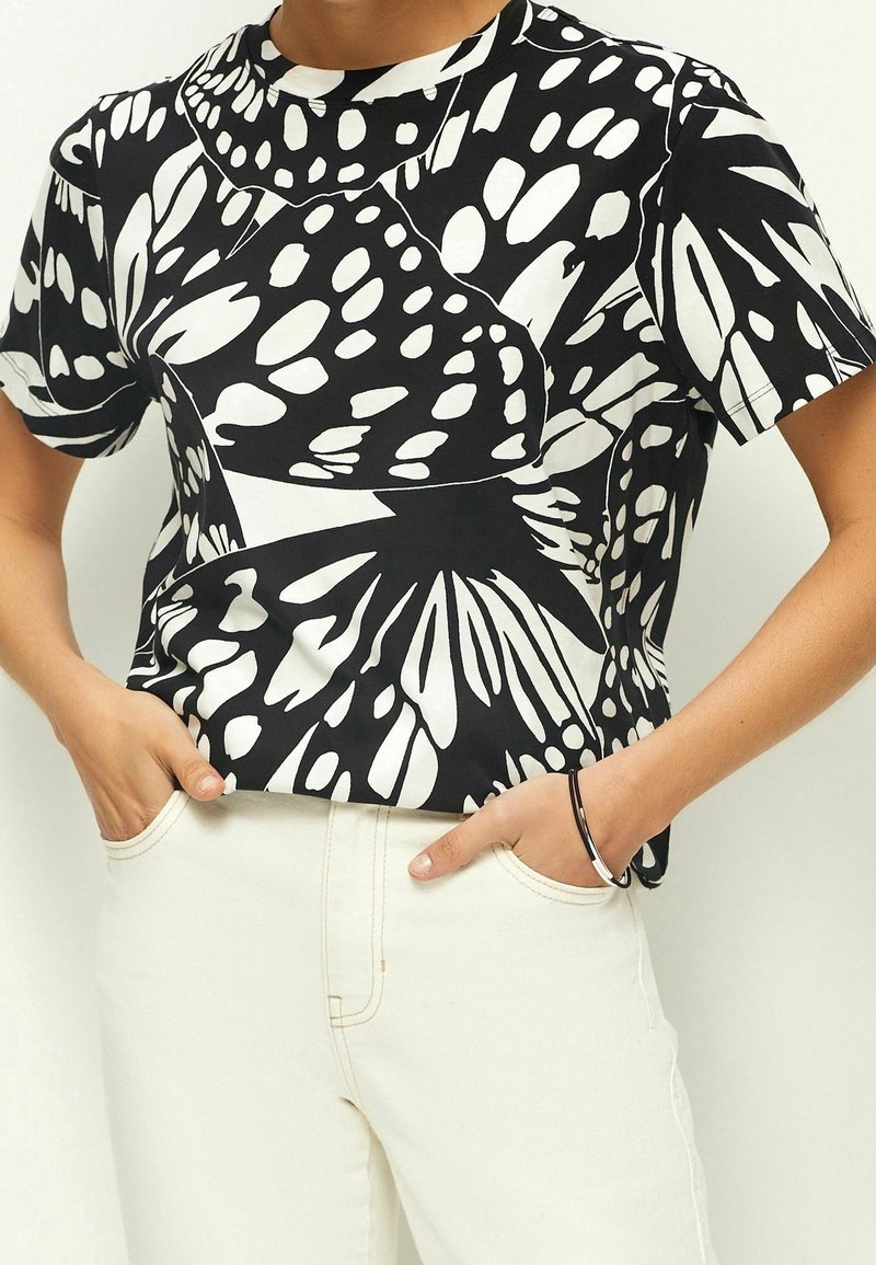 Personne portant un haut manches courtes à motif papillon noir et blanc, les mains dans les poches d’un pantalon blanc taille haute.