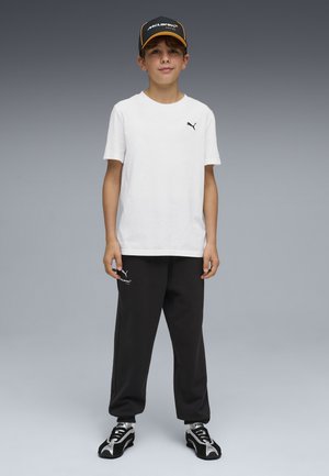 Niño de pie que lleva una camiseta blanca de Puma, pantalones negros McLaren Puma, zapatillas negras y blancas de Puma, y una gorra negra de McLaren.