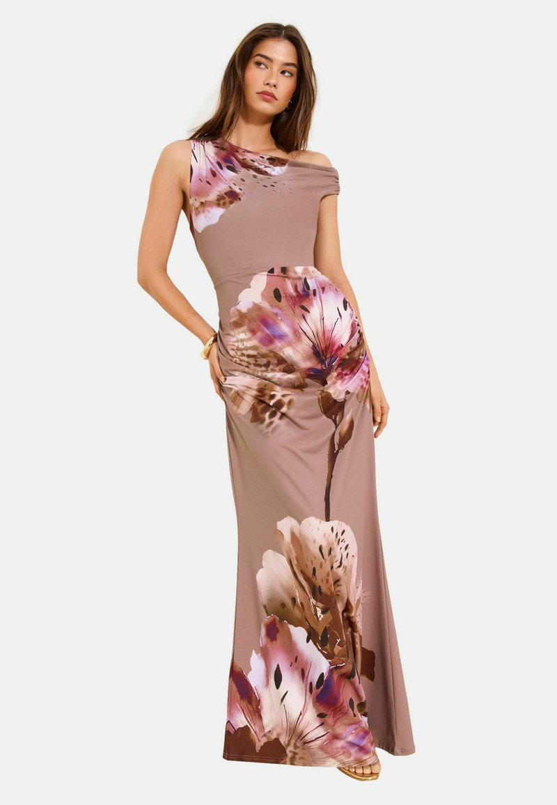 Frau in einem ärmellosen mauvefarbenen Maxikleid mit großen rosa und braunen Blumenmustern, die mit einer Hand auf der Hüfte und der anderen locker posiert.