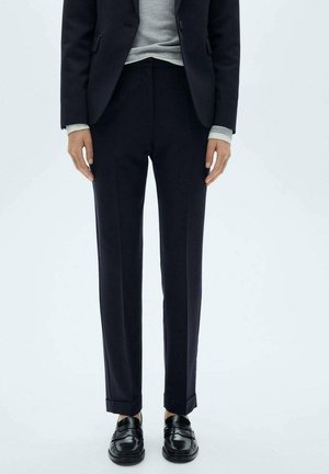 Person, die schwarze, maßgeschneiderte Hosen, einen schwarzen Blazer, einen grauen Pullover und schwarze Leder-Loafer trägt, steht vor einem hellen Hintergrund.