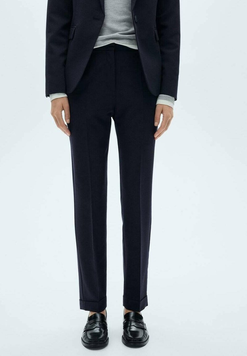 Personne portant un pantalon noir sur mesure, un blazer noir, un pull gris et des mocassins en cuir noir, debout devant un fond clair.