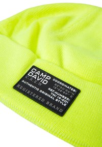 Neon yellow knit beanie featuring a black fabric label with white text, including brand name and coordinates. Soft texture and snug design.