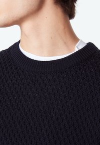 Pull en maille bleu marine avec un motif texturé, col rond, et une chemise blanche à superposer en dessous, mettant en valeur le contraste des matériaux.