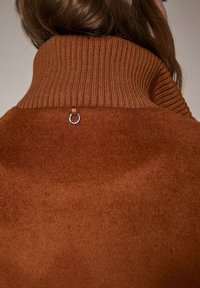 Prenda marrón con cuello alto y acanalado, con una textura suave y un detalle de anillo plateado en la parte posterior. El tejido parece suave y cálido.