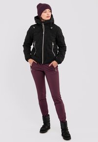 Schwarze isolierte Jacke mit strukturiertem Muster, Frontreißverschlüssen und Kapuze. Kombiniert mit bordeauxfarbenen Slim-Fit-Hosen mit hexagonalen Akzenten und schwarzen Stiefeln.