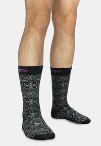 Schwarze Socken mit weißen geometrischen Mustern, gerippten Bündchen und kleinen Flaggenakzenten. Hergestellt aus einem weichen, strukturierten Stoff.