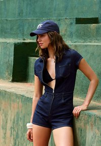 Jeans kombinezon, temno modra barva, kratki rokavi, zadrga spredaj, podrobnosti žepov, oblečen preko črnega bikinija, v kombinaciji z mornarsko modrim klobukom. Strukturirana zelena ozadja.