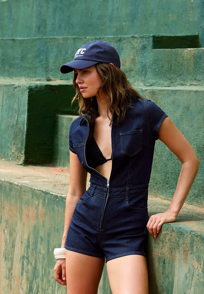 Jeans kombinezon, temno modra barva, kratki rokavi, zadrga spredaj, podrobnosti žepov, oblečen preko črnega bikinija, v kombinaciji z mornarsko modrim klobukom. Strukturirana zelena ozadja.