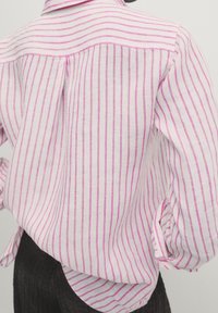 Vue arrière d'une personne portant une chemise blanche à rayures verticales roses et un pantalon noir texturé, manches de la chemise retroussées.