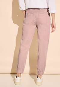 Light pink tapered pants with elastic cuffs, pockets, and a soft texture, paired with white sneakers against a neutral background.