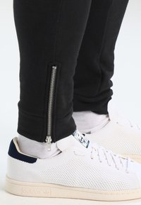 Svarta sweatpants med sidodetalj med dragkedja, tillsammans med vita perforerade sneakers. Byxorna har en slim fit och elastiska mudd.