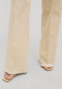 Beige wijde broek met rafelende randen, gemaakt van lichtgewicht stof, gecombineerd met beige vierkante sandalen.