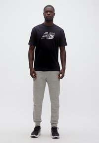 Άνδρας που στέκεται φορώντας μαύρο μπλουζάκι New Balance, γκρι φόρμα και μαύρα αθλητικά παπούτσια μπροστά σε λευκό φόντο.