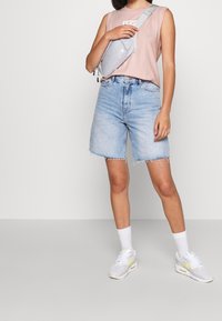 Camicetta senza maniche di un rosa chiaro, pantaloni corti in denim blu chiaro a vita alta con orlo sfrangiato, borsa a tracolla grigia e sneakers bianche con dettagli in neon.