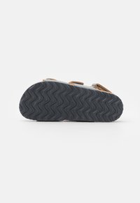 Grå sandal med bruna accenter, med en strukturerad svart gummisula och justerbara remmar. Sulan har ett vågigt mönster för bra grepp.