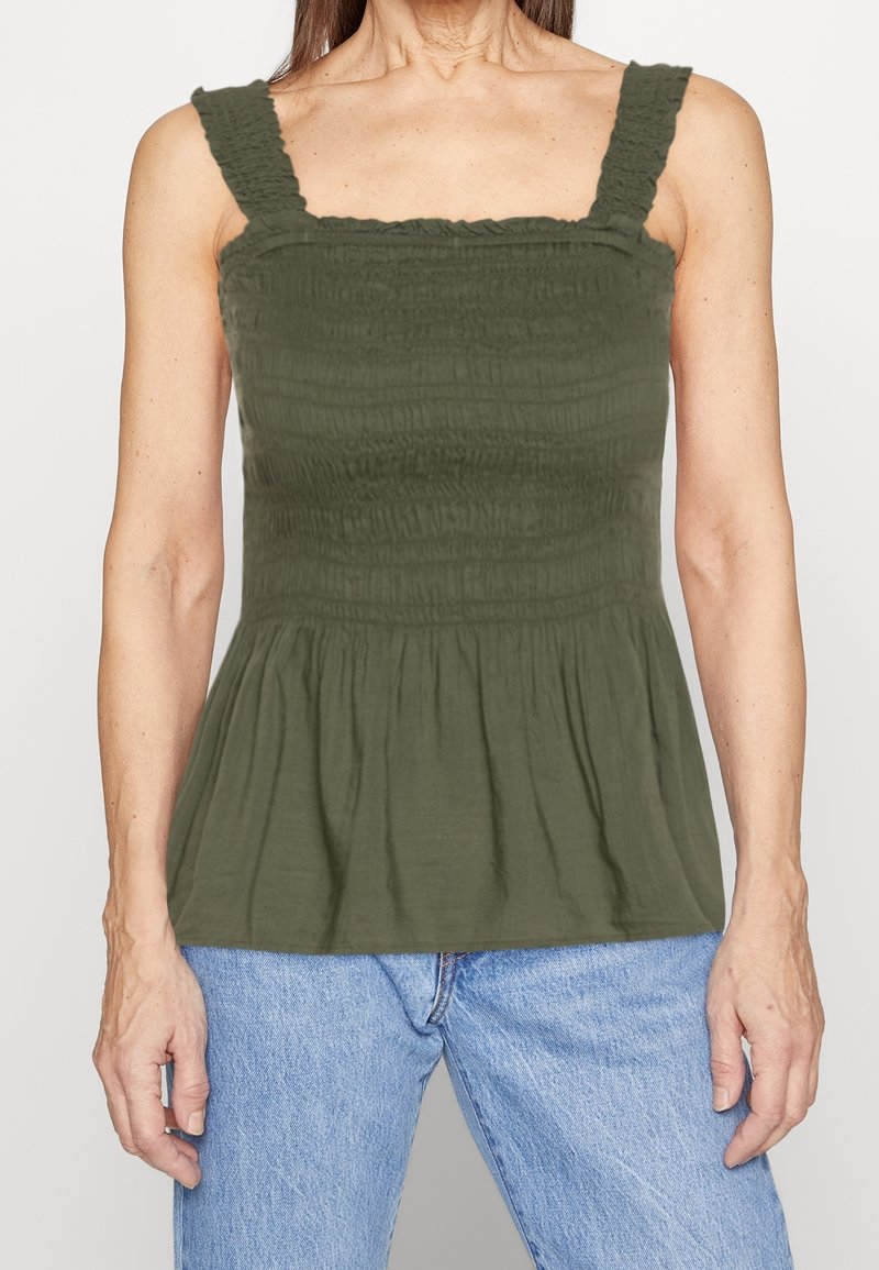 Haut débardeur smocké vert avec encolure carrée et bretelles volantées, associé à un jean bleu clair. Tissu texturé et détail d'ourlet à peplum.