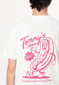 White t-shirt featuring a pink graphic of a smiling hot dog character, with "Tommy's Diner" and "24/7" text in playful fonts.