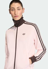 Svetlo roza jopica z zadrgo in rjavim visokim ovratnikom, z temno rjavimi poudarki in tremi belimi črtami na vsakem rami. Adidas logotip na prsih.