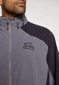 Γκρίζο αδιάβροχο με μαύρες λεπτομέρειες, πλήρης φερμουάρ και ρυθμιζόμενος γιακάς. Διαθέτει κεντητό λογότυπο σε μαύρο χρώμα στο στήθος. Ελαφρύ ύφασμα.