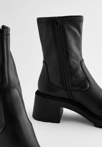 Botas de tobillo de cuero negro con cremallera lateral, tacón cuadrado y suela texturizada. Presentan líneas limpias y detalles de costura mínimos.
