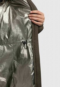 Cappotto verde oliva con fodera riflettente argento. Presenta un cordoncino regolabile e una texture trapuntata lungo i bordi.