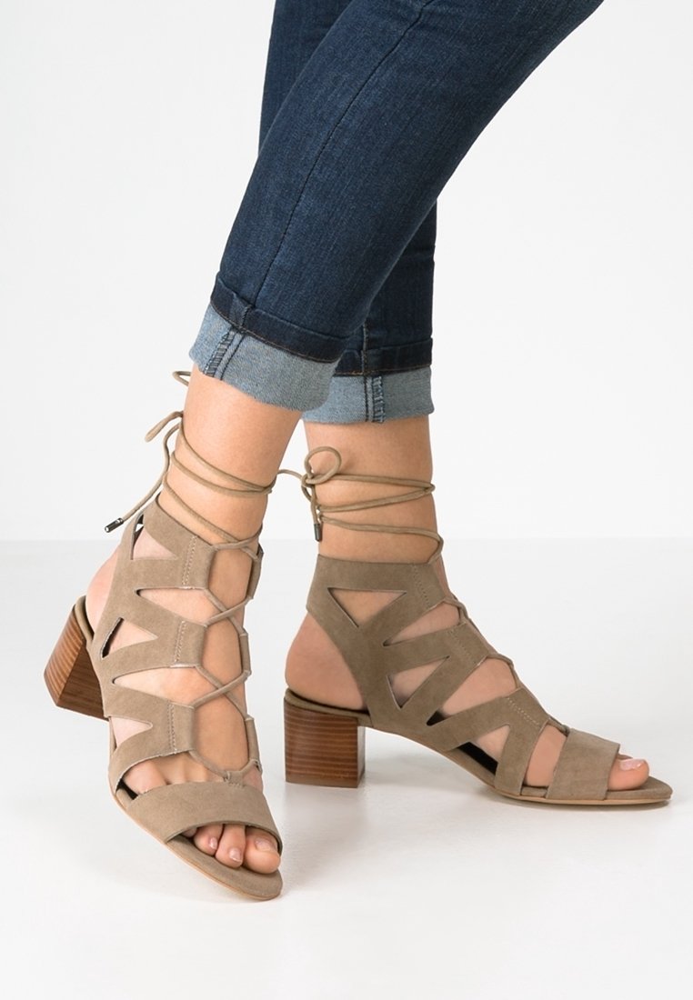 Sandales à talons en suede beige avec découpes triangulaires, design lacé et talon en bois empilé. Portées avec un jean foncé retroussé.