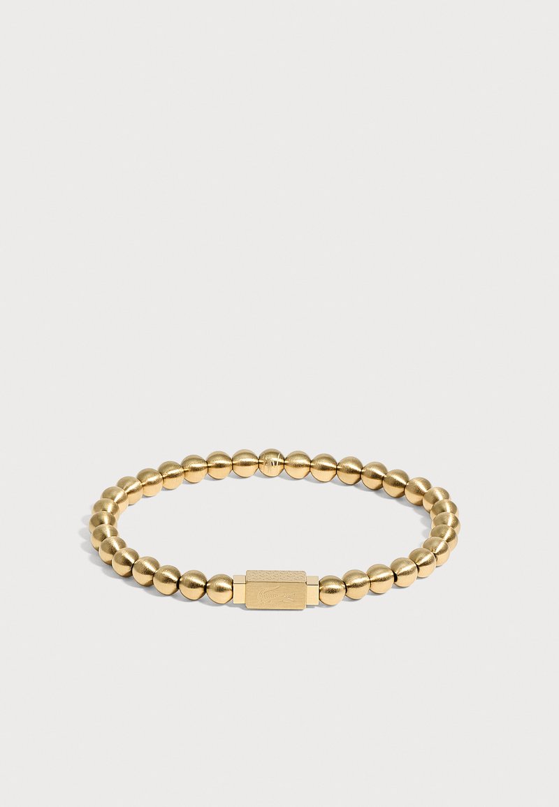 Bracelet en perles dorées avec fermoir rectangulaire présentant un design gravé subtil, montré sur un fond clair uni.