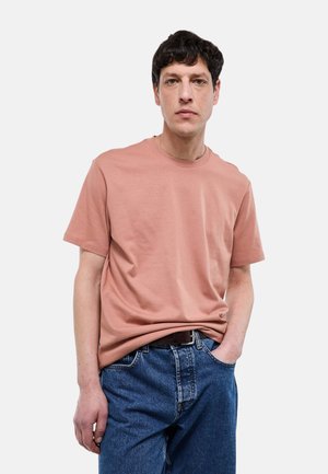 Homme aux cheveux courts et sombres portant un t-shirt rose poudré uni, rentré dans un jean bleu, debout avec une main dans la poche.