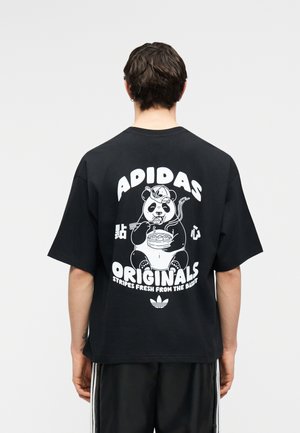 Osoba nosící černé tričko Adidas s bílým obrázkem pandy, která jí nudle, a nápisem „ADIDAS ORIGINALS“ na zádech.