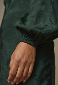 Sötétzöld szövet virágmintával, összegyűrt mandzsettákkal és sima, enyhén textúrált felülettel. A kéz a fókuszban, természetes bőrszínnel.