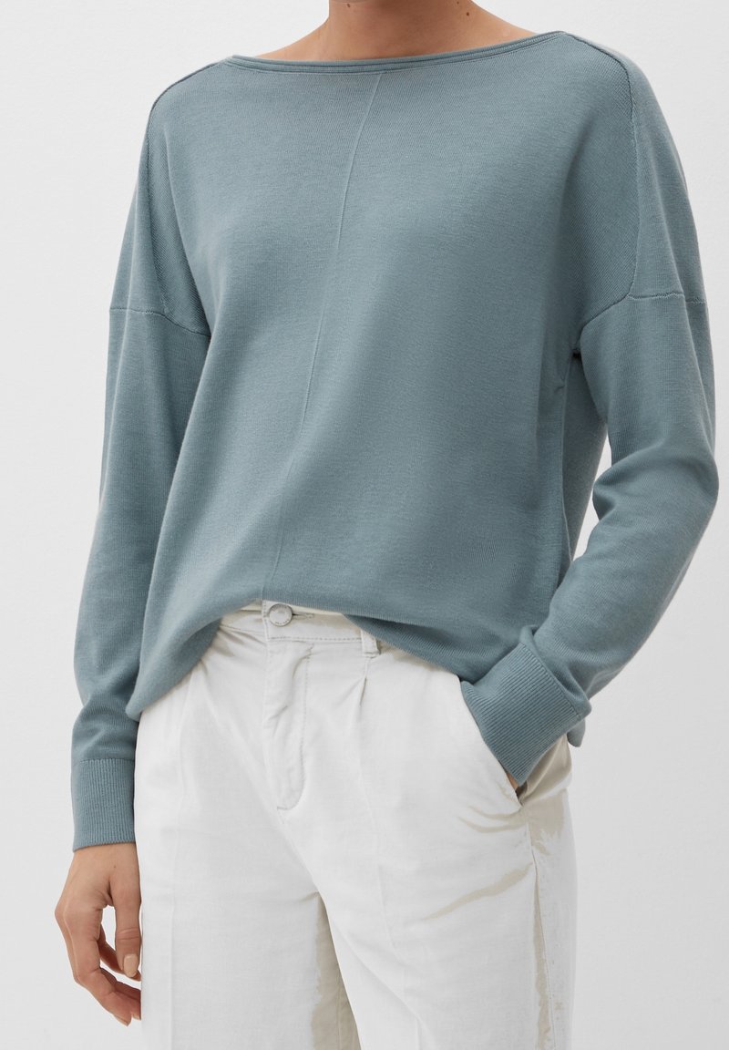 Personne portant un pull à manches longues bleu clair rentré dans un pantalon blanc taille haute, avec une main dans la poche et un fond neutre.