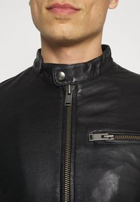 Svart läderjacka med hög krage, framzipper och silverfärgade detaljer. Ytan är slät med synliga sömnadsdetaljer.