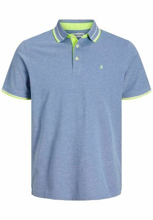 Homme portant un polo bleu clair avec une bordure vert néon et un jean bleu, debout devant un fond blanc uni.