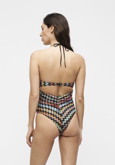 Γυναίκα με πολύχρωμο ολόσωμο μαγιό με ζικ-ζακ μοτίβο, δετό στον λαιμό με φιόγκο και άνοιγμα στην πλάτη, εμφανίζεται από πίσω σε απλό φόντο.