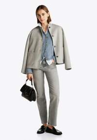 Cape en laine grise avec fermeture à bouton, superposé à un gilet en tricot bleu, associé à un jean gris et des chaussures en cuir noires, tenant un sac à main noir.