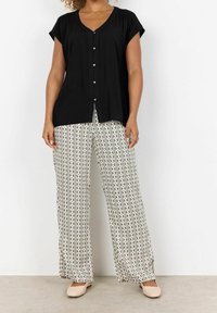 Chemise noire boutonnée à manches courtes, associée à un pantalon large à motifs crème et noir avec des dessins géométriques. Chaussures aux tons neutres.